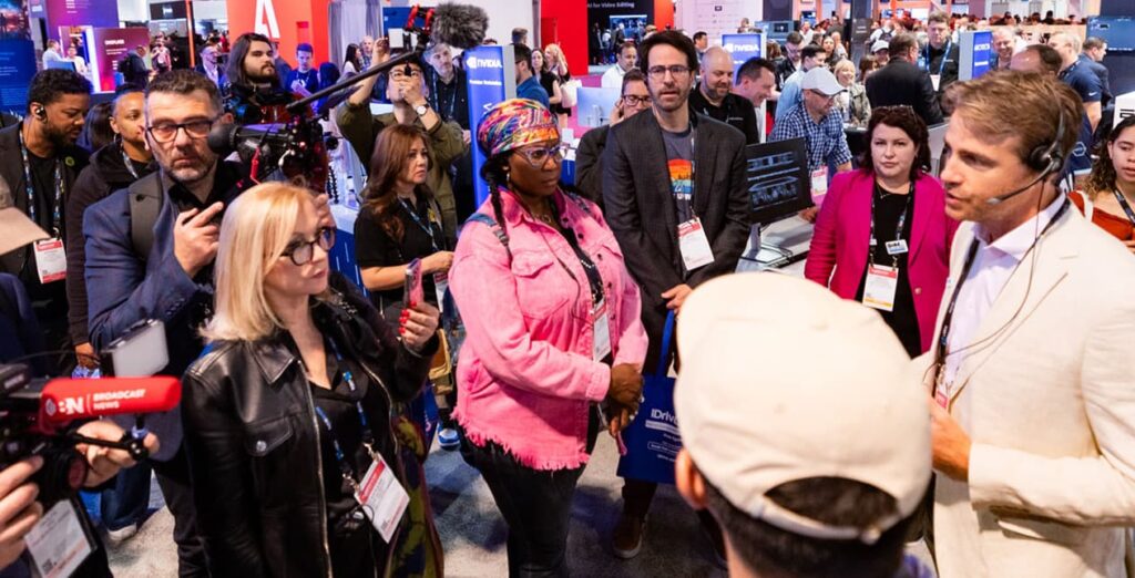 Photo of people on the Show floor with cameras, looking at a man speaking on the right.